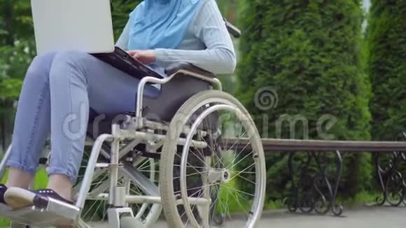 一位戴着传统围巾残疾的阿拉伯青年妇女使用坐在轮椅上的笔记本电脑视频的预览图