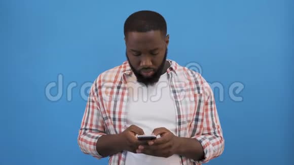 非洲男人被隔离在蓝色穿着白色连帽衫看着镜头竖起大拇指拿着智能手机感觉很开心客户视频的预览图