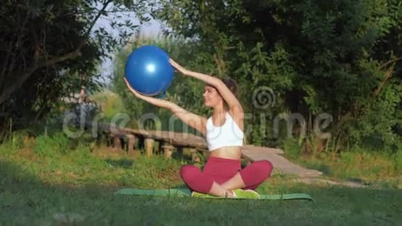 户外运动有魅力的女孩做手臂肌肉运动大健身球坐在草地上的垫子上视频的预览图