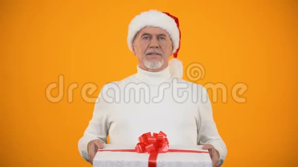 戴着圣诞老人礼帽的慈祥的老人在镜头前展示礼品盒和微笑的惊喜视频的预览图