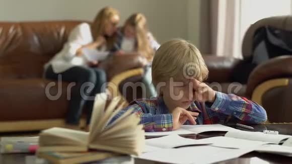 小男孩在家做作业时坐在桌子旁手里拿着笔记本母亲和妹妹坐在后面视频的预览图