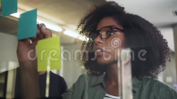 年轻的女商人和她的同事在玻璃上用黏贴纸写信视频的预览图