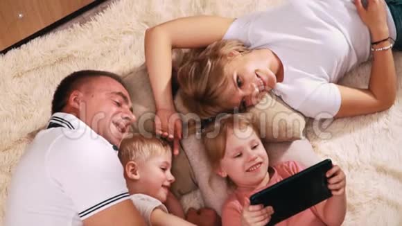 一家人带着两个小孩子躺在温暖的毯子上微笑着看着平板电脑它抱着她的女儿爸爸视频的预览图