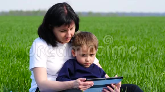 母子俩在公园里放松坐在绿草上家庭使用平板电脑播放看电影或制作视频的预览图