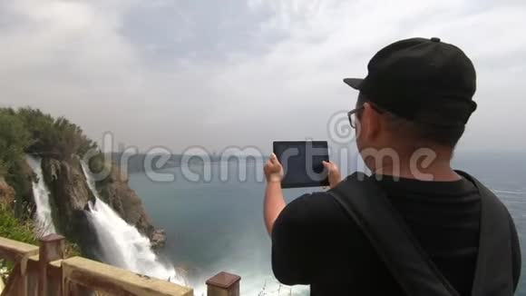 一位游客背着一个背包在陡峭的斜坡附近拍摄一个美丽的瀑布视频的预览图