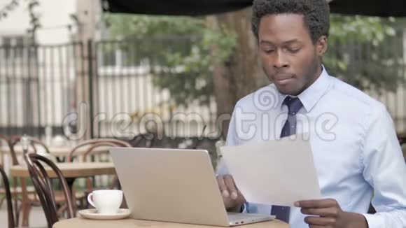 非洲商人阅读合同和在户外咖啡厅使用笔记本电脑视频的预览图