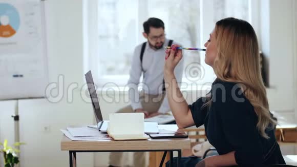 快乐年轻美丽的金发商务女性思考铅笔在现代轻办公室男性经理在背景视频的预览图