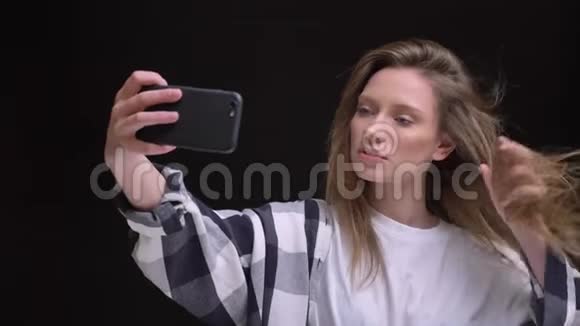 金发白种人长发女孩在黑色背景下在智能手机上制作自拍照片的肖像视频的预览图