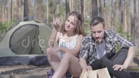 画像中的年轻人在森林里点燃一把火而可爱的年轻女人坐在附近可爱的情侣在户外休息视频的预览图