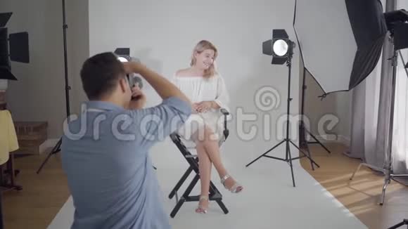 著名的专业摄影师为坐在白色背景椅子上的迷人优雅女人拍照视频的预览图