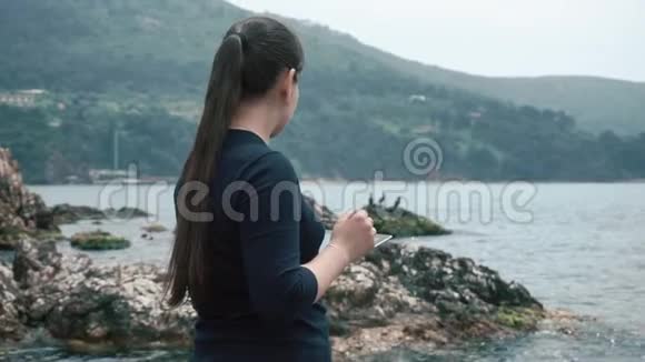 女游客站在岩石海滩上欣赏风景拿着平板电脑视频的预览图