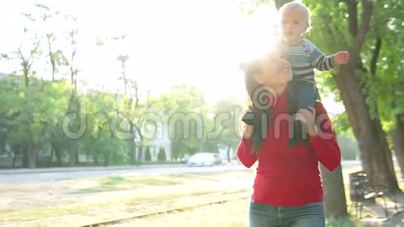 年轻的母亲和她的小儿子微笑着在公园里散步一个男孩坐在一个穿红色毛衣的女孩身上视频的预览图