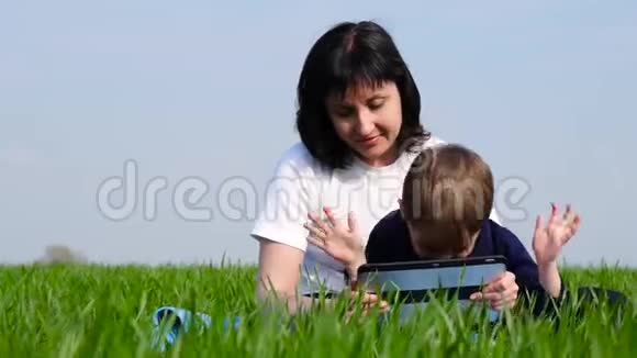 母子俩在公园里放松坐在绿草上家庭使用平板电脑播放看电影或制作视频的预览图