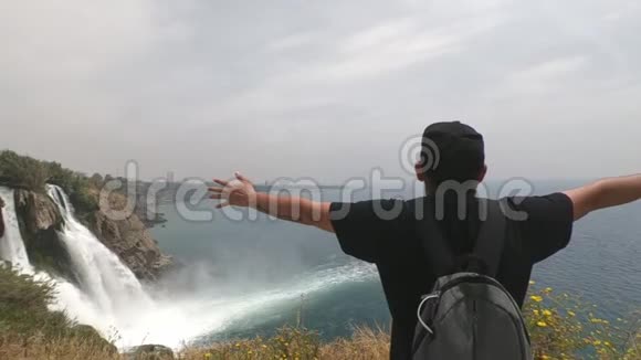 游客背着一个背包站立的手臂伸到不同的方向靠近陡峭的瀑布视频的预览图
