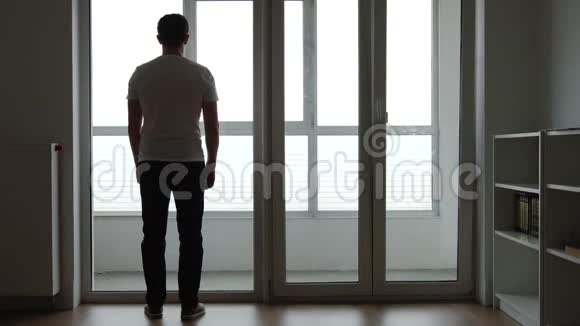 悲伤的年轻人站在家的一扇大窗户附近的剪影视频的预览图