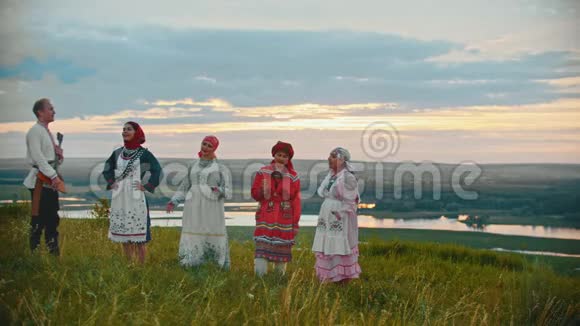 穿着俄罗斯传统服装的年轻人站在河流和岛屿的背景下的田野上视频的预览图