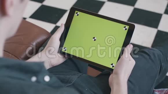 一位身穿黑色衬衫的年轻人坐在家里的沙发上看着带有彩色钥匙绿色屏幕的平板电脑视频的预览图