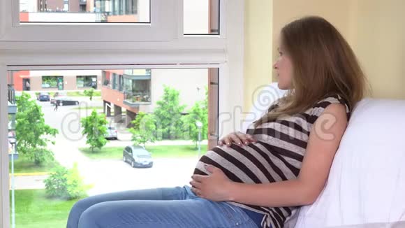 年轻的孕妇抚摸着温柔的胃透过窗户看着视频的预览图