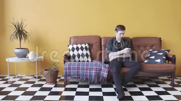 穿着黑色衬衫和牛仔裤的年轻人坐在棕色的皮革沙发上拿着智能手机库存录像周末视频的预览图