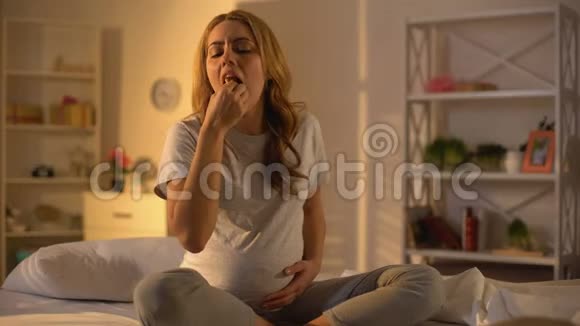 期待女性吃蛋糕抚摸坐在床上的腹部怀孕的虚弱视频的预览图