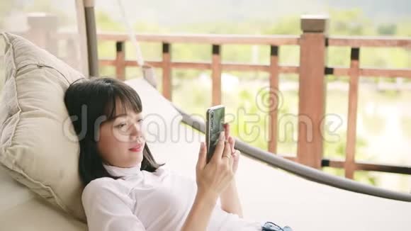 年轻的亚洲女人牙牙咧嘴幸福地微笑着手里拿着智能手机轻松的生活方式视频的预览图