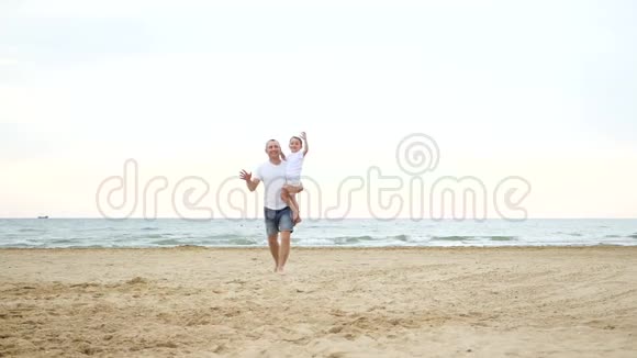 一个男人带着一个孩子在他的肩膀上沿着靠近海边的沙滩缓慢地运动视频的预览图