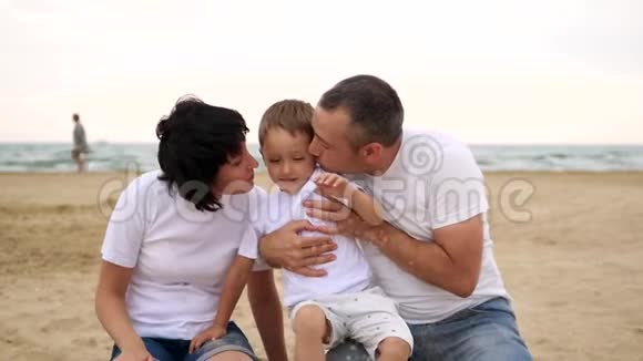 快乐的爸爸妈妈在海边的沙滩上亲吻孩子微笑着慢吞吞地视频的预览图