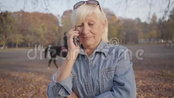 成熟的欧洲女人站在秋天公园用智能手机说话的肖像积极的高白种人女士视频的预览图
