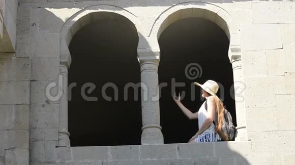 戴着背包的帽子的年轻女人自拍坐在老窗的拱门里夏日阳光明媚旅费视频的预览图