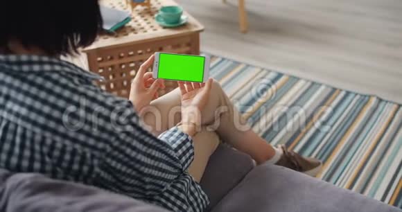 年轻女性在家坐在沙发上使用智能手机屏幕上有绿色的模拟屏幕视频的预览图