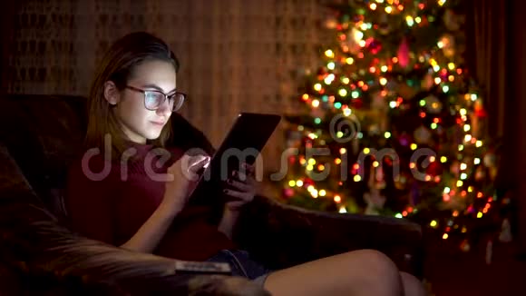 一个年轻的女人坐在圣诞树的背景上手里拿着一块平板电脑房间是装饰和填充的视频的预览图
