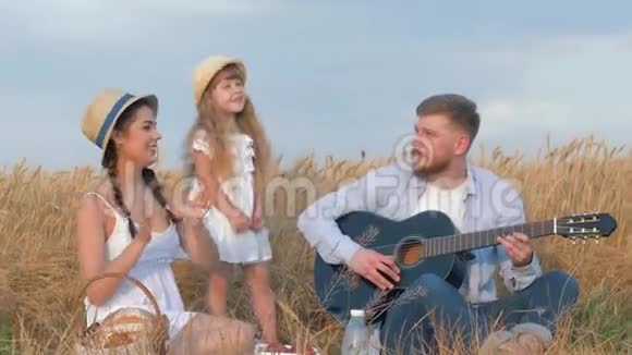 家庭户外娱乐年轻人弹吉他而他的女人和小可爱的女儿戴着草帽和白色的视频的预览图