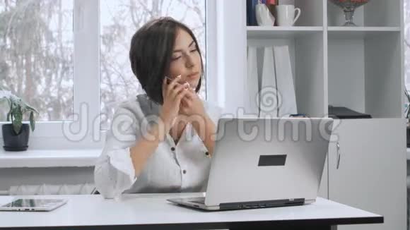 穿着白色衬衫的女人在手机上说话在他的笔记本电脑上工作在办公室工作的女商人视频的预览图