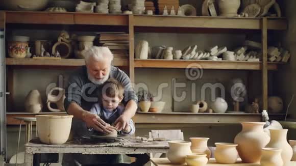 爱爷爷的专业陶工正在教他的小孙子在陶工车轮上塑造粘土小男孩视频的预览图