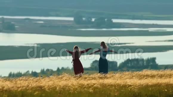 在阳光明媚的日子里两个年轻的女人伸出手臂在田野上旋转湖的背景上视频的预览图