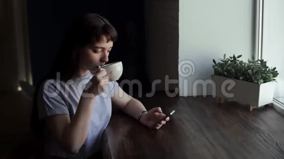 女孩在电话上写字在窗户附近的咖啡馆喝咖啡或茶视频的预览图