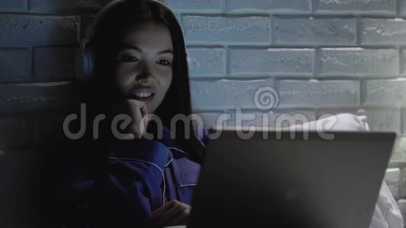 在夜间用笔记本电脑观看喜剧节目的耳机中微笑女性放松视频的预览图