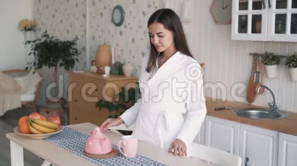 穿着白色浴袍的女孩在家厨房早晨用杯子倒茶视频的预览图