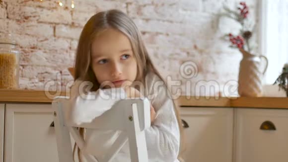 一个忧郁的小女孩蓝眼睛金发穿着白色连衣裙和毛衣坐在阁楼风格的房间里的椅子上视频的预览图