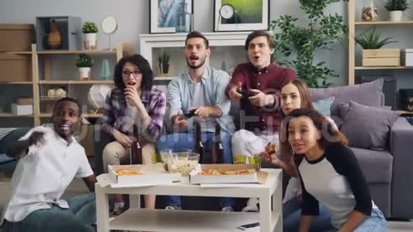 年轻的快乐朋友玩电子游戏说着吃着玩着在家聚会视频的预览图