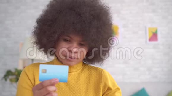 漂亮的非裔美国女孩发型是非洲式的手里拿着银行卡现代公寓里拿着笔记本电脑视频的预览图