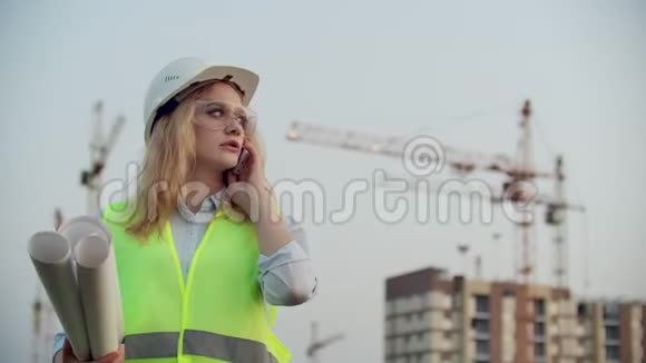 会说话的女人戴着头盔在电话里背景是施工手里拿着起重机图纸女性视频的预览图