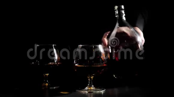 豪华白兰地穿着黑色衬衫的人在黑色背景上戴着两个玻璃杯附近的一瓶金色干邑白兰地白兰地视频的预览图