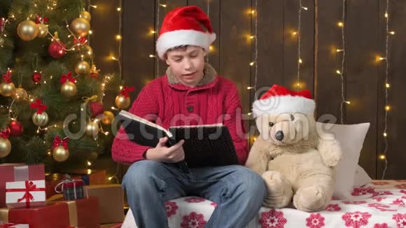 圣诞老人帮手男孩看书熊玩具坐在室内靠近装饰圣诞树灯圣诞快乐和节日快乐视频的预览图