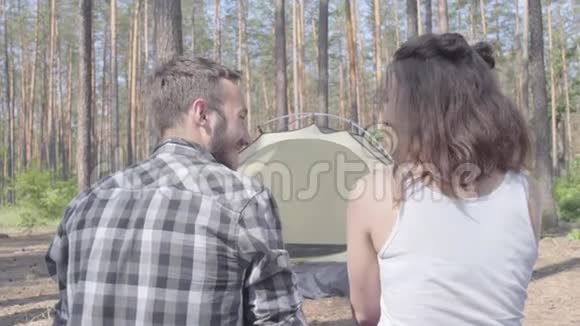 在森林里年轻的男人和女人背对着镜头坐在一起背景上的帐篷那个人视频的预览图