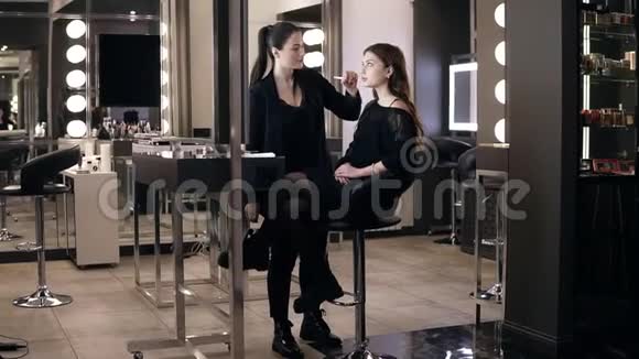美丽的少女穿着休闲黑色连衣裙坐在镜子前的椅子上而女化妆师则在施妆视频的预览图