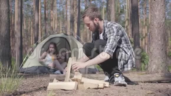 穿着格子衬衫的帅哥在户外准备木柴生火女孩坐在帐篷里玩四弦琴视频的预览图