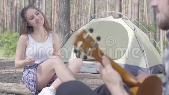 英俊的长胡子的年轻人在帐篷里玩四弦琴而漂亮的年轻快乐的女人坐在他面前爱视频的预览图
