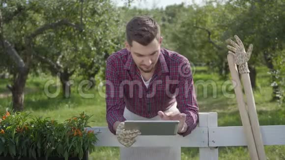 年轻的农民用平板电脑站在篱笆后面的前景夏天的花园在背景那个人视频的预览图