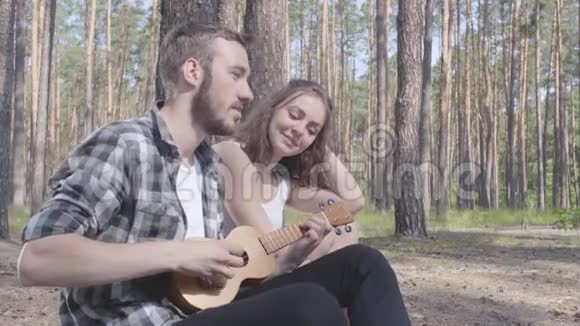 肖像英俊的留胡子的年轻人玩四弦琴而漂亮的年轻快乐的女人坐在附近可爱的一对视频的预览图